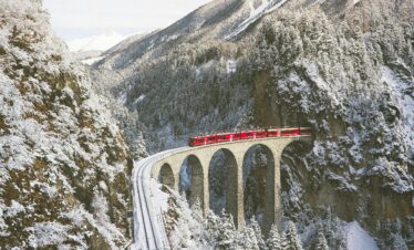 Bernina Express crossing the famous Landwasser Viaduct, surrounded by snow-covered mountains in the Swiss Alps.