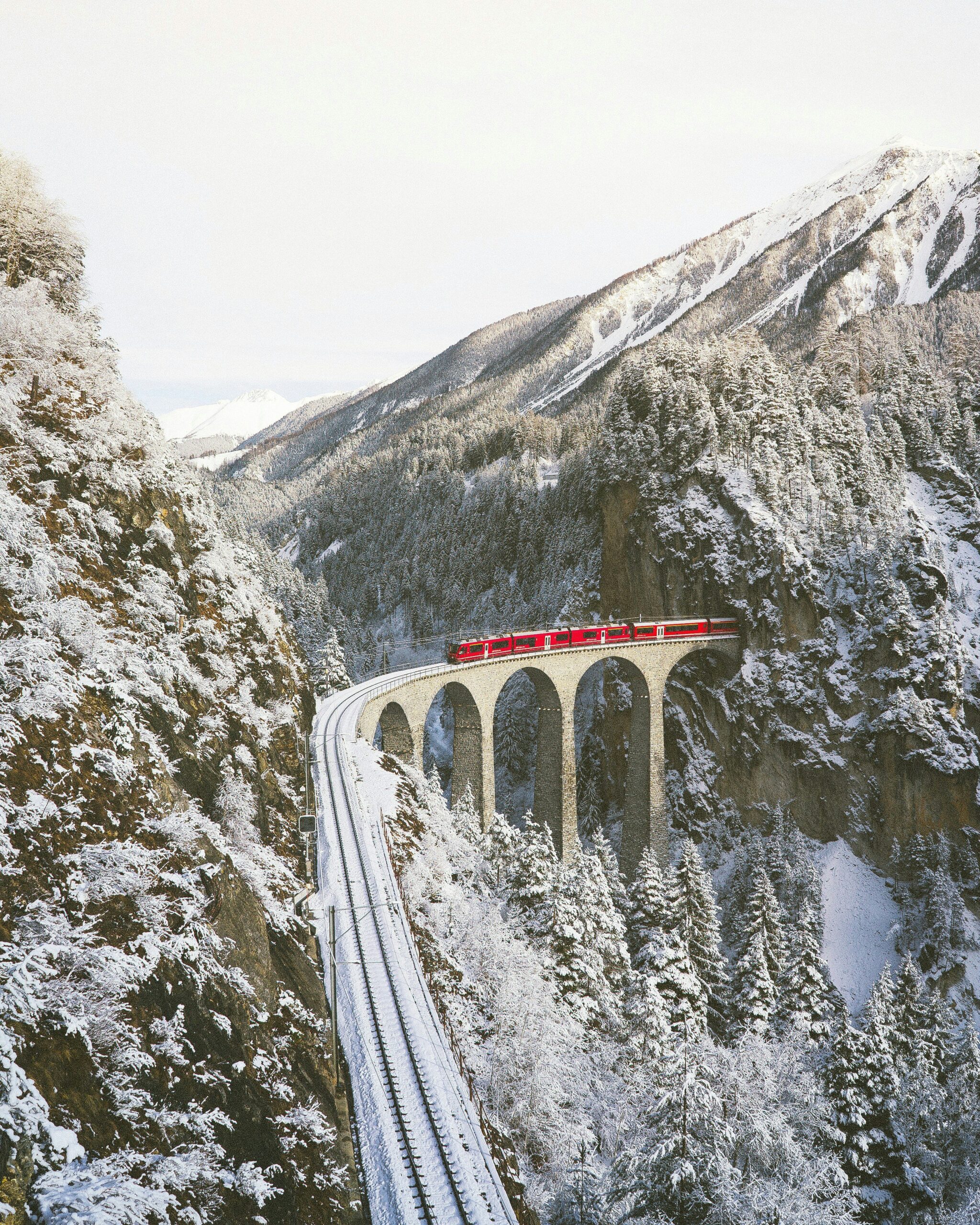 Bernina Express crossing the famous Landwasser Viaduct, surrounded by snow-covered mountains in the Swiss Alps.