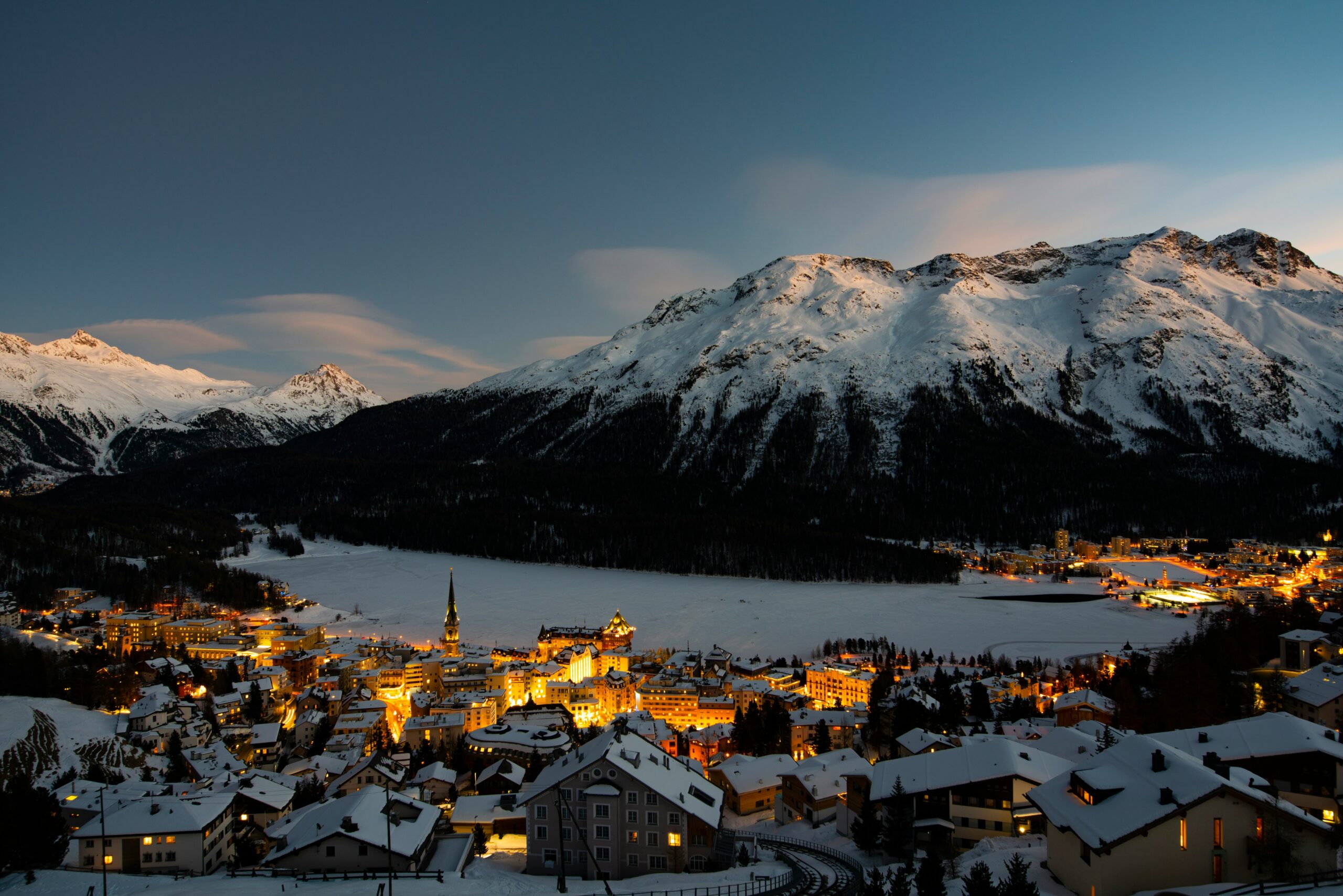 St. Moritz at almost night, with a frozen lake reflecting the fading light and snow-covered mountains in the background.