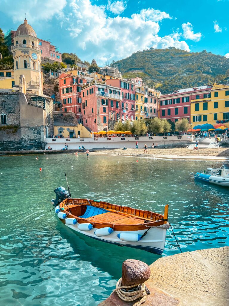 Boat floating on a serene lake in Italy, with colorful, traditional Italian houses along the shore, featuring vibrant yellows, oranges, and reds, set against lush green hills.