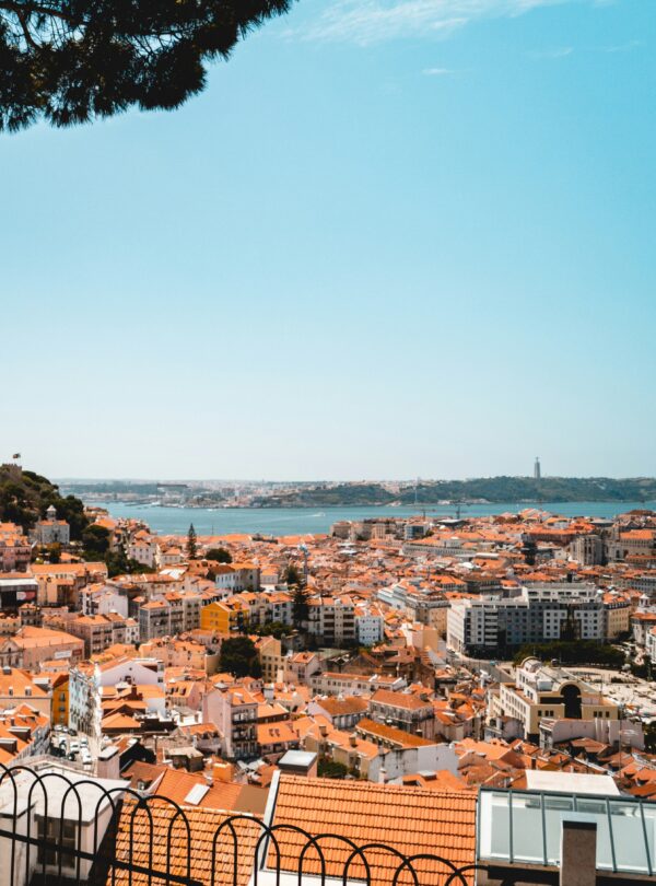 A panoramic view of Lisbon, featuring the iconic Castelo de São Jorge, the Cristo Rei statue, and the Ponte 25 de Abril (25th of April Bridge) in the distance, framed by the city’s charming rooftops.